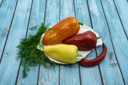 There are three bell peppers on a white plate; red, orange, yellow. On the table next to it is red...
