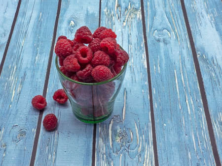 A transparent glass cup filled with ripe raspberries. Several berries fell onto the blue painted...