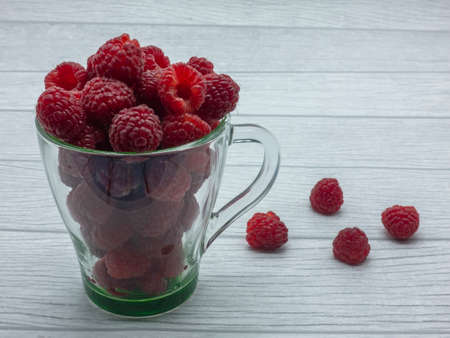 Transparent glass mug filled with ripe raspberries. Several berries are lying side by side on the table. White background.の写真素材