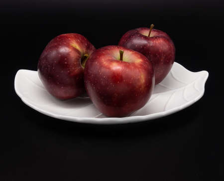 On a black background, a white plate in the form of a leaf. There are three bright fresh red apples on it. Contrasts.の写真素材