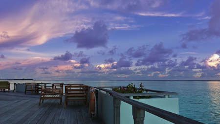 There are tables and chairs on a wooden deck in the middle of the ocean. Calm turquoise water. There...