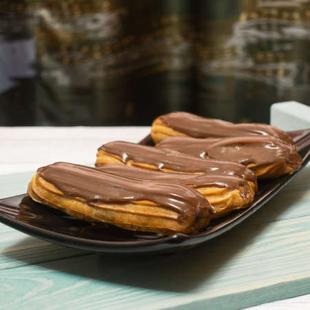 Five fresh eclairs covered with chocolate glaze lie on a rectangular platter. The plate is on a blue wooden tray. Soft background.の写真素材