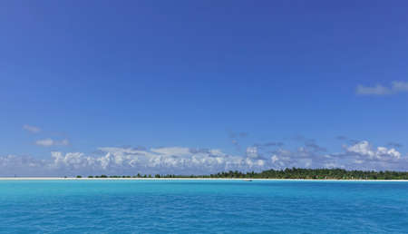 The calm ocean shimmers with aquamarine shades. An island with a sandy beach and palm trees is visible on the horizon. There are picturesque clouds in the azure sky. Maldives.の写真素材