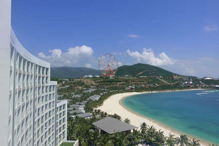 Top view of the turquoise sea, the curved line of the sandy beach, mountains. A fragment of a tall building, several villas, is visible. In the distance a colorful Ferris wheel. Vietnam. Nha Trang.のeditorial素材