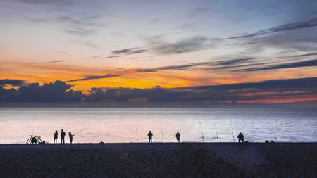 Evening twilight on the seashore. Silhouettes of fishermen, fishing rods, bicycles are visible. The sky is colored purple-orange. Black Sea. Russia. Sochiの写真素材
