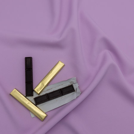 Four stick-shaped pieces of chocolate. Two are wrapped in golden paper. Lower left corner. Lilac...