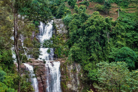 The waterfall cascades down from the rocks. The water is foaming. Lush green tropical vegetation surrounds it. Sri Lanka. Nuwara eliyaの写真素材