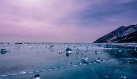 On the smooth frozen lake are scattered many shining shards of ice, similar to diamonds. Cracks and surface reflections. In the distance, snow-capped mountains. Pinkish sunset sky. Lake Baikal. Russiaの写真素材
