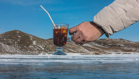 In a man's hand is a glass of mulled wine. Bright red wine, sliced fruit, plastic tube. Background - frozen lake, snow-capped mountains. Winter sunny day, Lake Baikalの写真素材