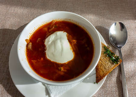 Lunch. Red Russian borscht with sour cream is already poured into the plate. On a saucer - a toast with herbs. There is a spoon on a beige cloth napkin. Top viewの写真素材