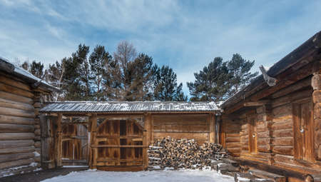 Inner village courtyard. The house, the fence - from natural unpainted logs. The gate is bolted. There is a stack of firewood against the wall. Background- coniferous trees against the blue sky.の写真素材