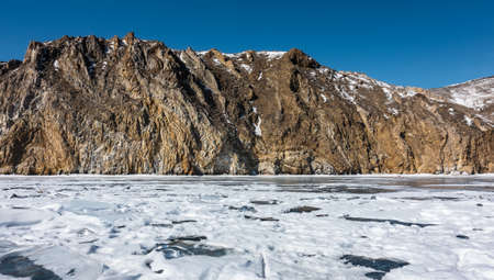 A picturesque mountain range, devoid of vegetation and covered with snow, rises above the icy lake. Rock with cracks and folds on the slopes. Stone texture. Clear blue sky. Baikalの写真素材