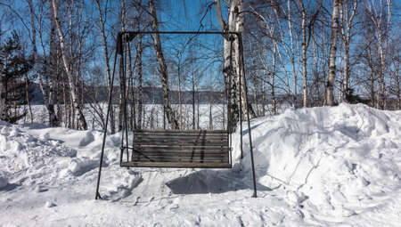 Wooden bench - swing on a metal frame, among the snowdrifts in the park. The seat is made of unpainted planks. Behind - a birch grove against the background of a bright blue sky, a mountain range.の写真素材
