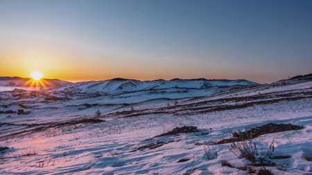 Sunrise over a snowy plain. The sun is over the ridge, its rays color the snow in golden hues. Dry grass and rare shrubs on the ground. The frozen Lake Baikal is visible.の写真素材