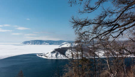 Top view of the border between the white ice of the frozen Lake Baikal and the blue water of the source of the non-freezing Angara River. Mountains against the sky. Bare tree branches in the foregrounの写真素材