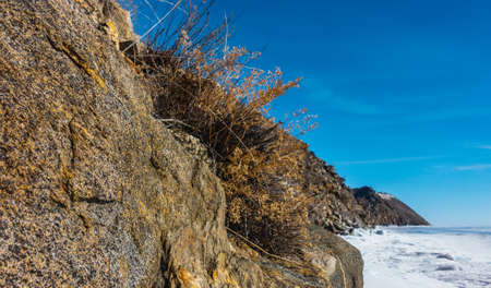 A slope of a granite rock on the shore of a frozen lake, against the background of a blue sky. Stone...