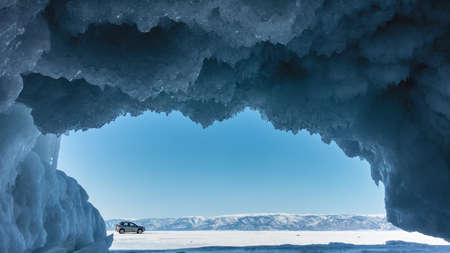 The vault and walls of the grotto in the rock are covered with blue icicles like lace frills. In the frame of the entrance, you can see a mountain range, the sky, a car on a frozen lake. Baikalの写真素材
