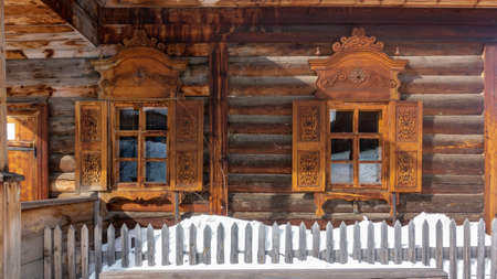 The old one-storey house was built of unpainted logs. There are shutters and carved decorations on...