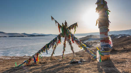 Wooden pillars are braided with multicolored ritual ribbons and placed in the form of a pyramid on the shore of a frozen lake. There is snow and dry grass on the ground. The sun is in the sky. Baikalの写真素材