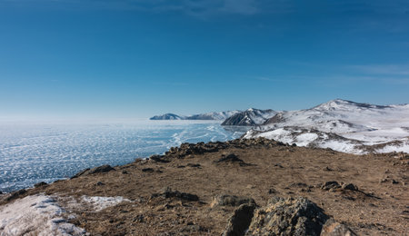 Winter Siberian landscape. Snow patterns are visible on the blue ice of the frozen lake. A mountain range on the background of an azure sky. Dry grass and rocks on the shore. Baikalの写真素材