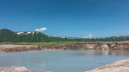 Thermal lake in the caldera of an extinct volcano. There are stones on the banks. Sun glare on the water. A plain covered with green grass. A mountain range against the blue sky. Kamchatka.の写真素材