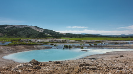 An amazing turquoise geothermal lake in the caldera of an extinct volcano. There is almost no vegetation on the stony soil. A mountain range against the blue sky. A summer day. Kamchatka. Uzonの写真素材