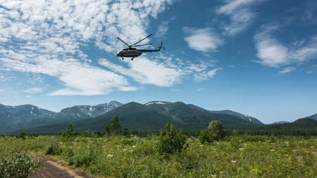 Grass and wildflowers grow on a green meadow. A dirt road goes to the mountains. A helicopter is flying against the background of a blue sky with clouds. Kamchatkaの写真素材
