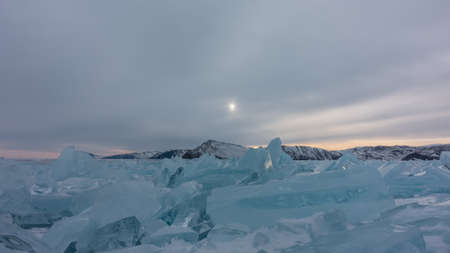 There is a block of turquoise hummocks on the surface of the frozen lake. Highlights on the edges. The setting sun in a cloudy sky. A mountain range in the distance. Baikalの写真素材