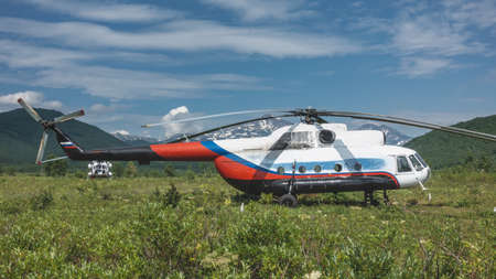 The helicopter is standing on a green meadow. Lush grass and wildflowers all around. Mountains on the background of a blue sky with clouds. Kamchatka Nalychevoの写真素材