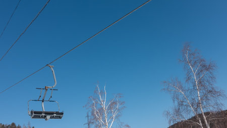 Chairlift. An empty seat is suspended on a cable. White trunks and bare branches of birch trees against a clear blue sky. Siberia. Listvyankaの写真素材