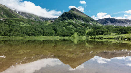 A picturesque mountain range on the shore of a lake. On the slopes there is green vegetation and areas of melted snow. A mirror reflection on calm water. Clouds in a blue sky. Kamchatka. Vachkazhetsの写真素材