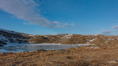 A small frozen lake is surrounded by hills. Snow on the ice and on the ground. In the distance, against the blue sky - the houses of the tourist base. Beautiful clouds. Siberia. Olkhon islandの写真素材