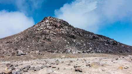 A rock devoid of vegetation, against a background of blue sky and clouds. Volcanic stones are scattered on the slopes and at the base. Kamchatka. Gorely volcanoの写真素材