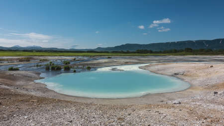 A small turquoise lake was formed from the hot springs. There are sulphurous deposits on the shores. A mountain range against a blue sky.  Kamchatka. The caldera of the extinct volcano Uzonの写真素材