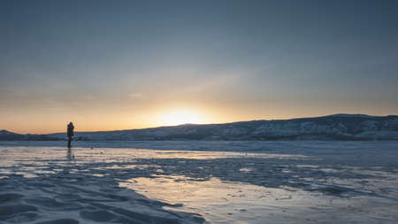 The setting sun turns the sky over the mountain range orange. There is a lonely man standing on a...
