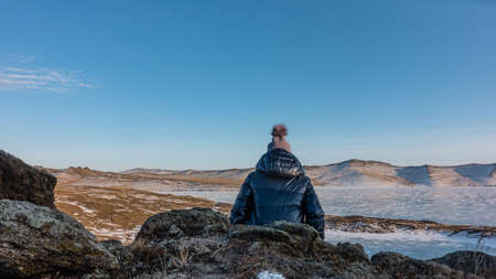A girl in a down jacket and hat is sitting on a granite rock. View from the back. In front of her is a frozen lake with snow patterns on the ice. A mountain range against a blue sky. Baikalの写真素材
