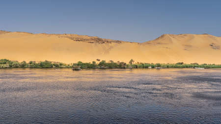 The Nile River flows calmly. Ripples on the water. There is green vegetation on the shore. High sand...