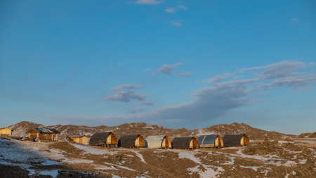 The small houses of the tourist base stand in a row on a hill. There are patches of snow on the surrounding ground. Picturesque clouds in the azure sky. Evening golden hour. Siberia. Olkhon islandの写真素材