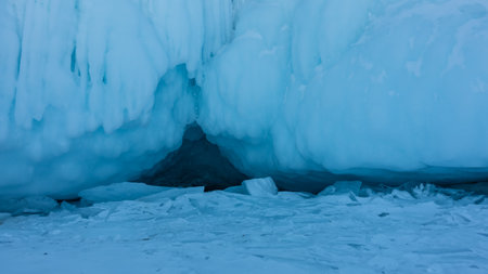 The base of the rock is covered with a thick layer of icicles and splashes. The entrance to the dark cave is visible. Fragments of ice on the surface of a frozen lake. Baikal.の写真素材