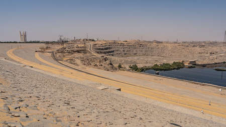 Aswan Dam in Egypt. Sandy-rocky soil on the banks of the Nile, technical buildings, power lines. Reflection on the smooth blue surface of the river.の写真素材