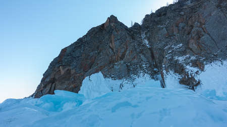 A picturesque granite rock against a blue sky. Cracks on the stone slopes. At the base is a block of turquoise ice hummocks. Lake Baikal.の写真素材
