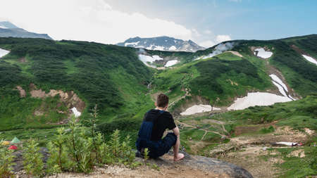 A man sits on a rock and looks down. There are tourist tents in the valley, people are walking along the paths. There is green vegetation and snow on the mountains. Steam from hot springs is visible.の写真素材