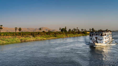 A cruise ship is sailing on the blue river. On the shore there is green vegetation, palm trees. High...