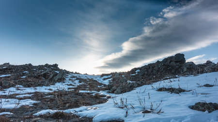 A hill against a background of blue sky and clouds. Snow on the ground. Dry grass among rocks and in snowdrifts. Siberiaの写真素材