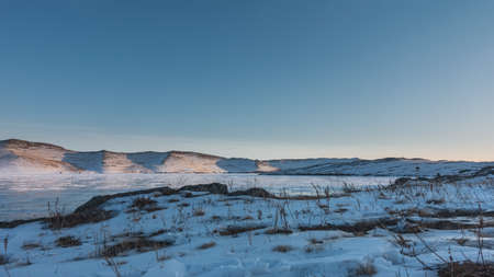 Winter in Siberia. There is snow and dry grass on the shore of a frozen lake. The sun illuminates...