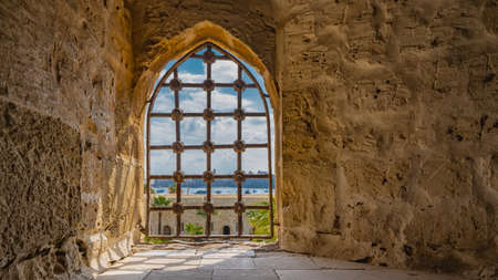 The window opening in the ancient Citadel of Qaitbay is barred. The texture of rough stone walls is visible. Shadows on the tiled floor. In the distance - blue sky, silhouettes of buildings in Egypt.の写真素材