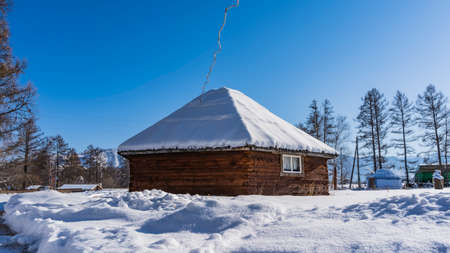 The old wooden ail house stands in the valley. Snow on the roof and on the ground. Bare trees...