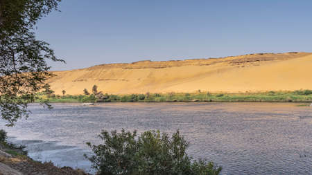 Green vegetation is visible on the banks of the Nile. A high sand dune against the blue sky. Ripples on the water. Egypt. Aswanの写真素材