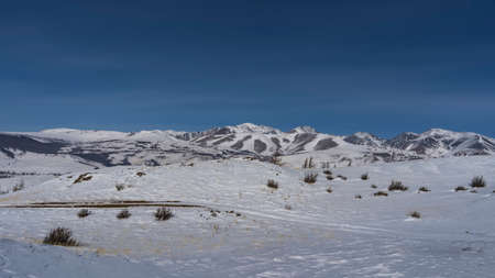A dirt road runs through a snow-covered valley. Yellow dry grass and bare shrubs are visible on the...