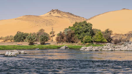 Boulders and green vegetation are visible on the banks of the Nile. Ripples on the blue water. Overturned boats lie on the sand. high dunes against the blue sky. Egyptの写真素材